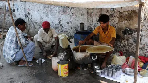 AFP via Getty Images Three men sit under a makeshift roadside canopy beside a worn wall, preparing and eating food at a small outdoor stall. A young man in a yellow shirt stirs a large metal pot placed over a stove, while metal cups, plates and containers are placed on the ground. 
